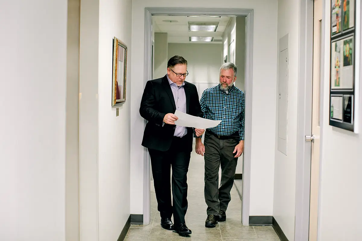 Two men walk down a narrow office hallway while looking at a large sheet of paper
