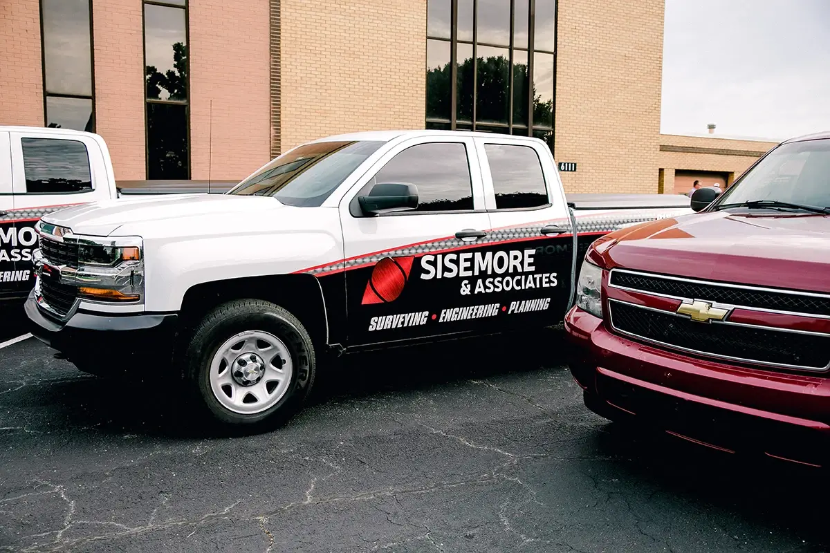 A white Sisemore & Associates pickup truck parked in a lot features black and red side graphics with "Surveying, Engineering, Planning" text under the logo.